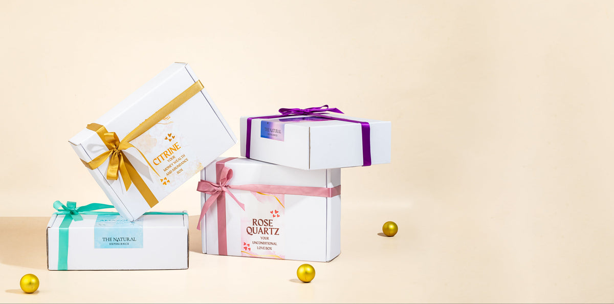 Four white gift boxes with colorful ribbons and printed labels are arranged on a warm beige studio background with small gold decorative spheres. Visible text includes CITRINE, ROSE QUARTZ, and THE SWEETNESS.